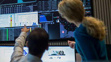 A man and woman in a control room and the man is pointing at one particular screen